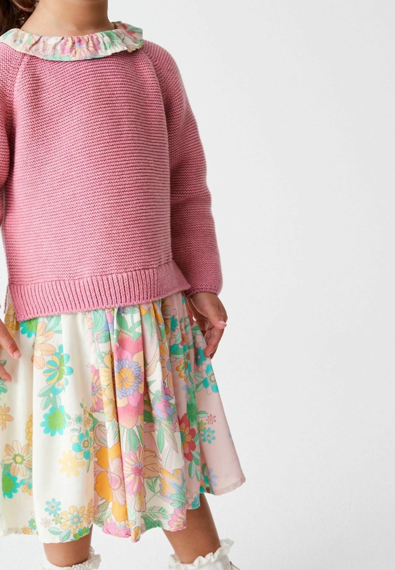 Vaaleanpunainen neulepaita, jossa on röyhelömäinen kukallinen kaulus, yhdistettynä polvipituiseen hameeseen, jossa on värikkäitä kukkakuoseja pehmeällä taustalla.