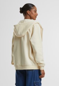 Crèmekleurige hoodie met een grote capuchon, zacht textiel, ontspannen pasvorm en ribgebreide manchetten. Zichtbare ritssluiting en stikseldetails.