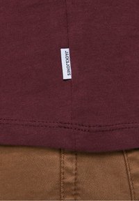 Gros plan sur un tissu bordeaux avec une étiquette "JACK & JONES" au-dessus de la couture des pantalons texturés marron.