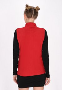 Vesta roșie din fleece cu guler înalt, având o textură netedă. Poartă deasupra unei bluze negre cu mânecă lungă, fără modele sau accesorii vizibile.