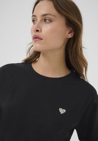 T-shirt en coton noir avec un col rond et un petit design de cœur orné de strass sur la poitrine.
