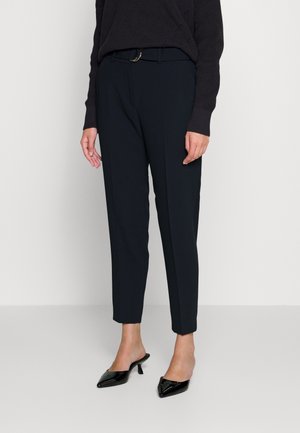 Pantalon ajusté bleu marine avec une texture lisse, doté d'une ceinture avec une boucle ronde, porté avec des mules à talons noirs.