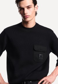 Giovane uomo in camicia nera con tasca a pattina e logo, in piedi con una mano sui fianchi contro uno sfondo bianco.