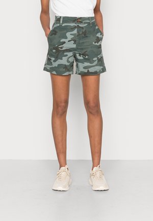 Personne portant un short camouflage vert et gris avec des poches, des baskets blanches, et un t-shirt blanc, debout les mains dans les poches.