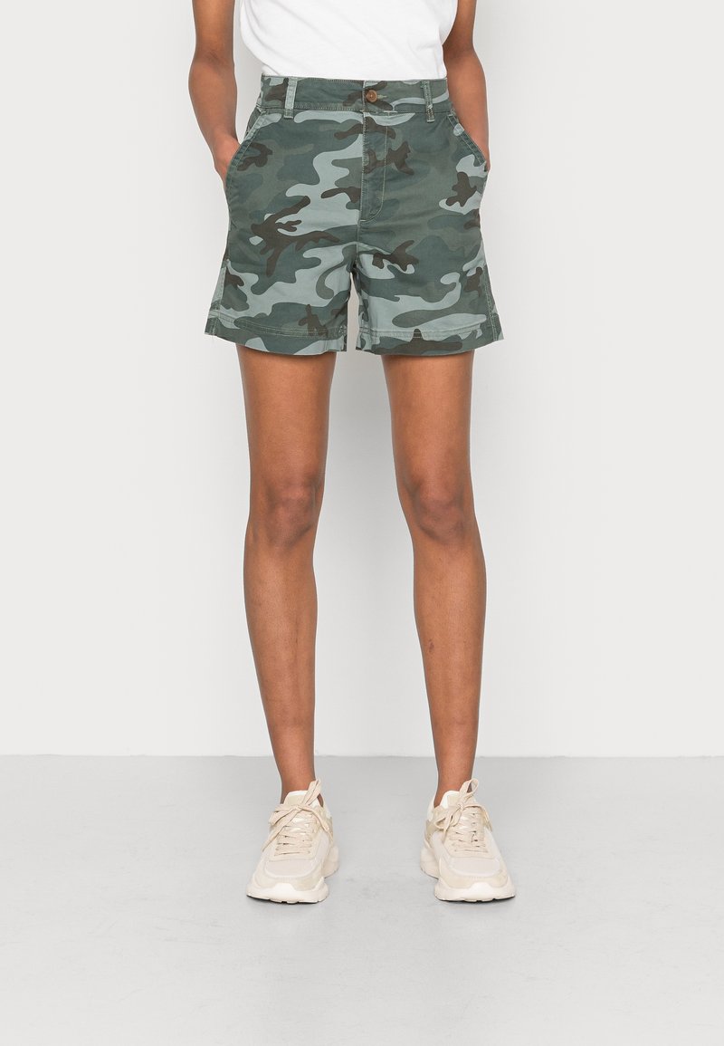 Personne portant un short camouflage vert et gris avec des poches, des baskets blanches, et un t-shirt blanc, debout les mains dans les poches.