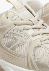 Beżowe buty sportowe wykonane z siateczki i materiałów syntetycznych, z amortyzującą podeszwą, zszywanym zapięciem oraz gumowymi akcentami w kolorze kremowym i metalicznego złota.