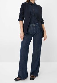 Žena v tmavě modrém blejzru, tmavě modré košili s knoflíky, širokých tmavě modrých džínách, černých síťovaných botách a černé crossbody tašce.