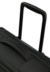 Valigia a rotelle nera con esterno in tessuto testurizzato, manico telescopico e impugnatura superiore robusta. Le cerniere sono visibili lungo le cuciture laterali.