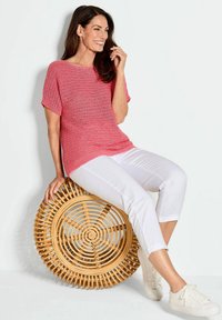 Roze gebreide top met textuur, korte mouwen, gecombineerd met witte cropped broek en witte sneakers. Zittend op een ronde bamboestoel.