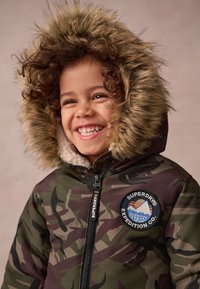 Kamelonīga pūkveida jaka ar mākslīgu kažokādu uz kapuces, ar rāvējslēdzēju un logo uzlīmi. Iezīmēta ar mīkstu oderi un tumši zaļu, brūnu un melnu krāsu paleti.