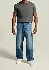 Homme portant un jean bleu ample, un t-shirt gris foncé à manches courtes et des chaussures blanches slip-on, debout devant un fond uni.