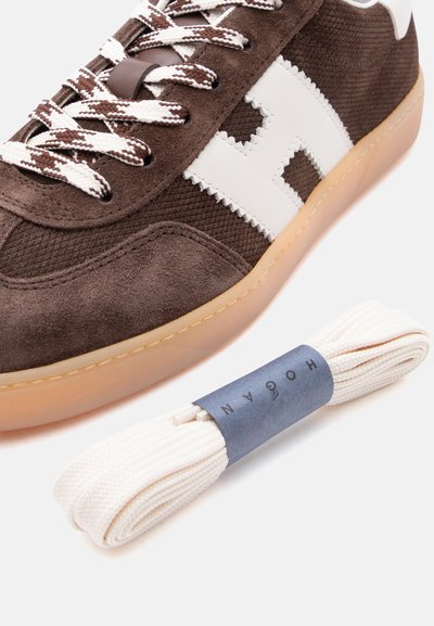 Καφέ σουέντ και πλεκτό αθλητικό παπούτσι με λευκό λογότυπο "H" και κορδόνια με σχέδιο, δίπλα σε ένα ζευγάρι τυλιγμένα λευκά ανταλλακτικά κορδόνια.