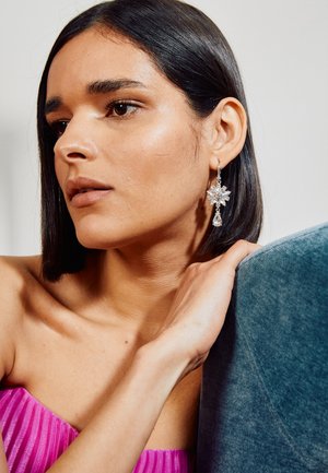 Woman with straight dark hair wearing a bright pink strapless top and large floral crystal drop earring resting hand on blue fabric.