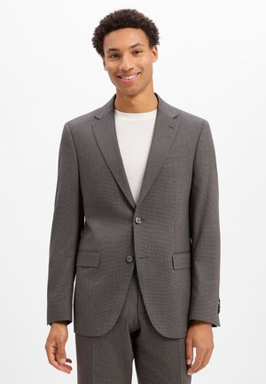 Man die een grijs colbert en broek draagt met een wit overhemd, glimlachend tegen een effen witte achtergrond.