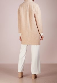 Cappotto beige morbido con colletto alto, maniche lunghe e polsini bianchi a coste; abbinato a pantaloni bianchi a gamba larga e scarpe con tacco beige.