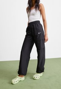 Zwarte cargobroek met een elastische tailleband, voorzien van twee zijzakken en een wit Nike-logo. Combinatie met lichtgroene klompen.