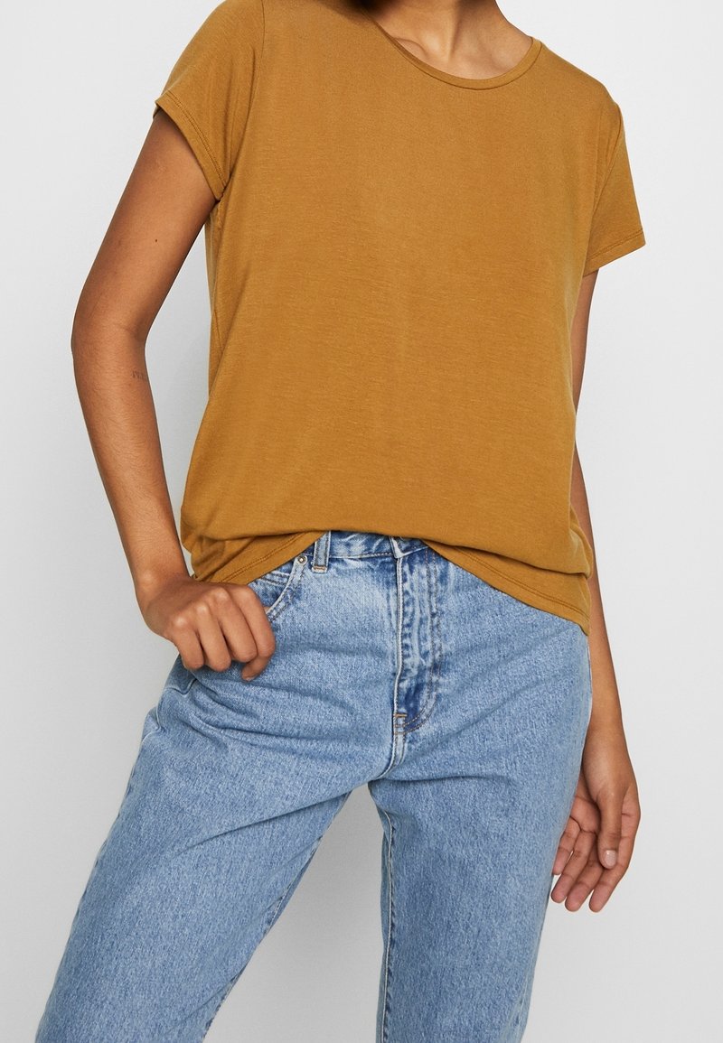 Femme portant un t-shirt à manches courtes jaune moutarde rentré dans un jean taille haute bleu clair, debout avec une main sur la hanche.