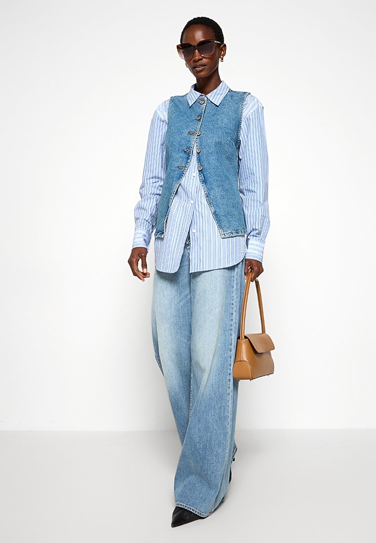 Denim-Weste über einem blau-weiß gestreiften Hemd, weite helle Denim-Hosen, braune Handtasche und schwarze spitz zulaufende Schuhe. Dunkle Sonnenbrille.