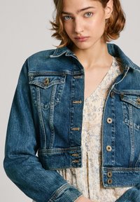 Chaqueta de denim en azul oscuro con diseño crop, dos bolsillos frontales, botones metálicos y costuras en contraste; llevada sobre un vestido floral.