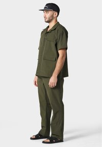 Camisa y pantalones de color verde oliva hechos de una mezcla de algodón; la camisa tiene mangas cortas, bolsillos frontales y cierre de botones; combinados con sandalias negras.