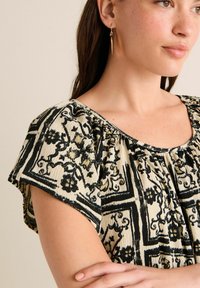 Pull crème avec des motifs floraux et géométriques noirs complexes, avec un col plissé et des manches courtes. Boucles d'oreilles pendantes dorées.