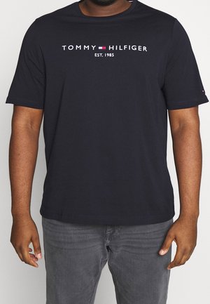 Uomo che indossa una maglietta nera di Tommy Hilfiger con logo e la scritta "EST. 1985", abbinata a jeans grigi scuri, in piedi contro uno sfondo bianco.