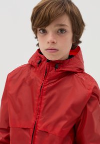 Giacca impermeabile rossa con zip frontale, cappuccio e coulisse regolabili; texture lucida e design minimalista senza motivi.