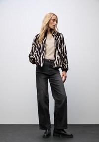 Veste bomber à motif zèbre noir et blanc avec une fermeture éclair, portée par-dessus un haut beige et assortie à un jean noir taille haute et des bottes noires.