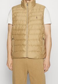 Beige, kviltad dunväst med hög krage, full frontal dragkedja och sidofickor. Har en liten logotyp på vänster bröstområde.