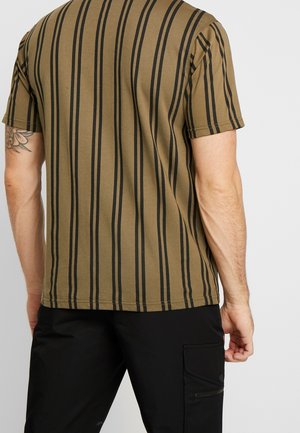 Olivengrøn T-shirt med vertikale sorte striber, korte ærmer og en lige kant. Synlig tatovering på armen; båret med sorte bukser.