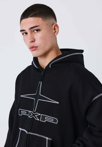 Sudadera negra con costuras blancas, cremallera frontal y un gran logotipo que dice "PXP" con un diseño de estrella en el pecho. Material suave, corte informal.