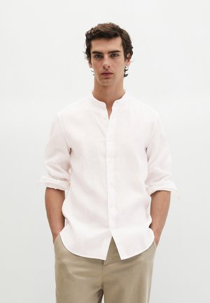 Młody mężczyzna o kręconych ciemnych włosach, ubrany w białą koszulę z kołnierzykiem mandaryńskim i beżowe spodnie, stojący z rękami w kieszeniach.