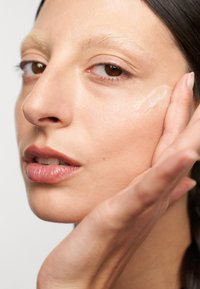 Close-up van een gezicht dat een crème aanbrengt; gladdere huid, natuurlijke make-up tinten, glanzende lippen en een heldere gelachtige product op de wang.
