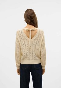 Maglione beige lavorato a maglia con scollatura a V profonda e dettaglio con fiocco, caratterizzato da un motivo testurizzato e una vestibilità ampia, abbinato a jeans scuri.