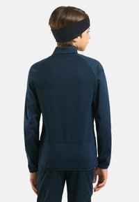 Junge in navyblauer Sportkleidung und schwarzem Stirnband, der von einer schlichten weißen Wand abgewandt ist.