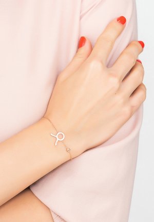 Rose gold bracelet featuring a glittering loop design with clear accents. Worn on a wrist with smooth skin and a soft pink fabric background.