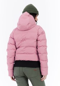 Růžová zimní bunda s kapucí, s prošívaným designem a černým žebrovaným lemem. Nosit s zelenými kalhotami a zelenou pletenou čepicí.