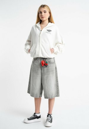 Młoda kobieta w białej bluzie z kapturem zapinanej na zamek z napisem, szarych szerokich szortach dżinsowych z czerwonym breloczkiem w kształcie serca oraz czarno-białych trampkach.