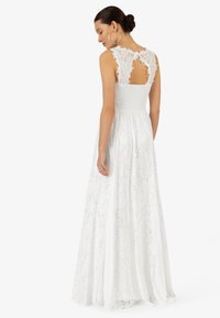 Robe en dentelle blanche avec un corsage smocké et un dos nu, ainsi qu'une longue jupe fluide ornée de motifs floraux délicats.