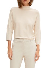 Maglione beige a maglia con collo alto e maniche a tre quarti. Texture liscia e dettagli a costine. Abbinato a pantaloni chiari e aderenti.