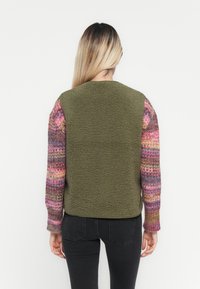 Fleece vest met een olijfgroene body en multicolore gebreide mouwen met roze, paarse en gele patronen. Getextureerde stof overal.