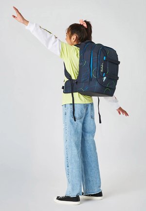 Kind mit großem blau-grün gemustertem Rucksack, steht mit ausgebreiteten Armen, trägt ein hellgrünes Hemd und blaue Jeans.