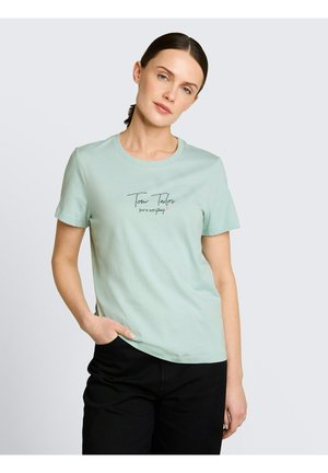 Vrouw met donker haar in een staart, gekleed in een lichtgroene shirt met korte mouwen met tekst en zwarte broek, tegen een effen lichtgrijze achtergrond.