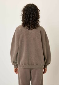 Prevelik rjav pulover s spuščenimi rameni, rebrastimi manšetami in robom. Mehek material, minimalno vidni šivi, nošen s usklajenimi hlačami.