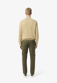 Maglione beige lavorato a maglia con colletto alto, abbinato a pantaloni verde scuro. Le scarpe sono bianche con dettagli verdi. È visibile un dettaglio in tessuto strutturato.