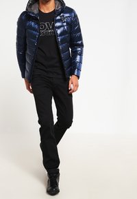 Marineblå pufferjakke med en blank finish, hætte og grå indvendig foring. Båret over en sort grafisk t-shirt og sorte bukser med sorte støvler.