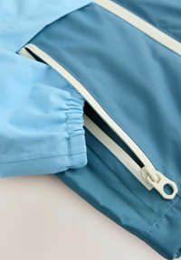 Giacca azzurro chiaro e teal con polsini elastici e dettagli in zip bianco. Tessuto liscio; design casual con un motivo a blocchi di colore.