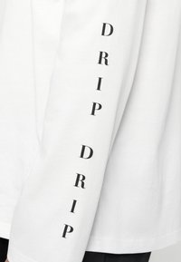 Άσπρο μακρυμάνικο πουκάμισο με μαύρο κάθετο κείμενο «DRIP DRIP» τυπωμένο στο μανίκι, μερικώς ορατό πάνω από σκούρο παντελόνι.
