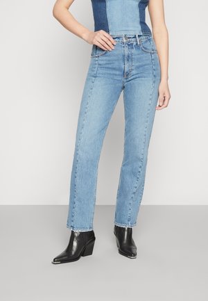 Femme portant un jean droit bleu clair et des bottines noires à bout pointu, debout avec une main sur la hanche.