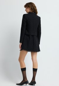 Sort tweedjakke med et cropped design, parret med en matchende nederdel med frynsekant. Accessoriseret med sorte strømpebukser og sko.